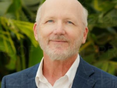 A portrait of San Diego Tourism Authority Content Manager James Hebert, who is wearing a blue coat and white shirt against a backdrop of greenery.