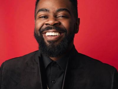 A portrait of the San Diego writer and community leader Ramel J. Wallace, who is smiling and wearing a black jacket and black shirt against a red backdrop.