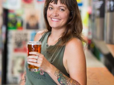 Writer Beth Demmon holds a glass of beer, smiling in a colorful brewery setting.