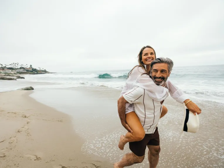 A joyful couple plays on the beach in San Diego, with the woman on the man's back, against a cloudy sky.