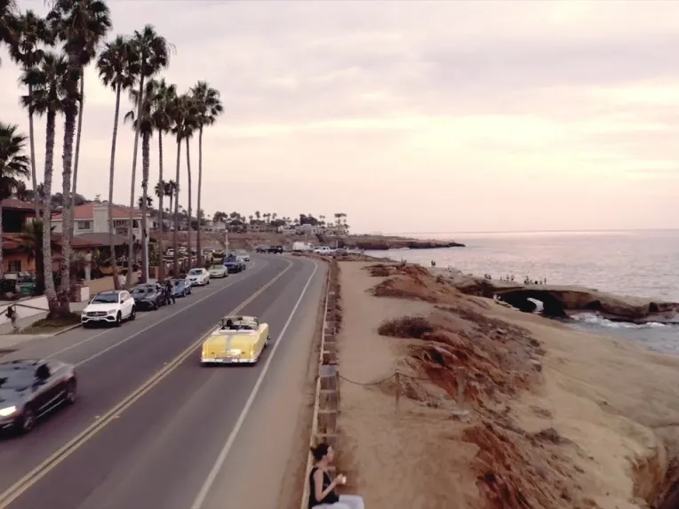 A yellow convertible drives along a coastal road lined with palm trees and houses, with ocean cliffs and parked cars on one side at sunset.