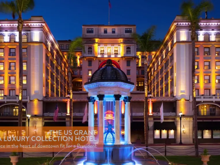 A brightly lit, grand hotel with a central fountain in front, viewed at night. The building features ornate architecture and palm trees flank the entrance.
