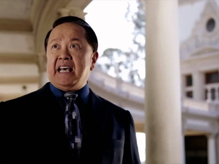 A man in a suit and tie speaks outdoors in front of a classical-style building with columns.