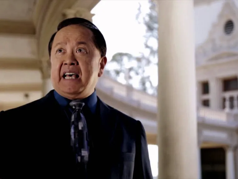 A man in a suit and tie speaks outdoors in front of a classical-style building with columns.