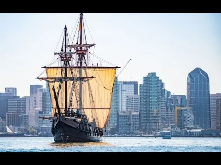 A large sailing ship with tan sails is on the water in front of a modern city skyline with tall buildings under a clear sky.