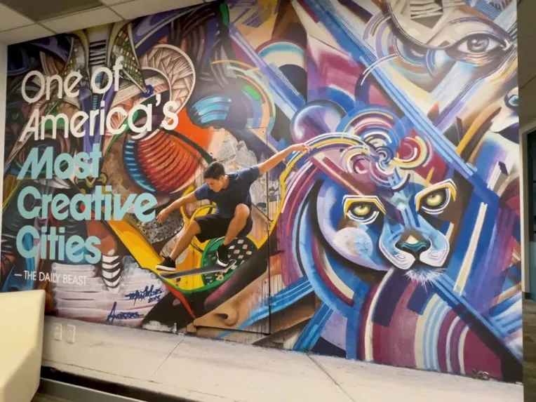 Colorful office mural with a skateboarder and vibrant animal motifs, labeled "One of America's Most Creative Cities, The Daily Beast." White sofa and framed certificates on left wall.