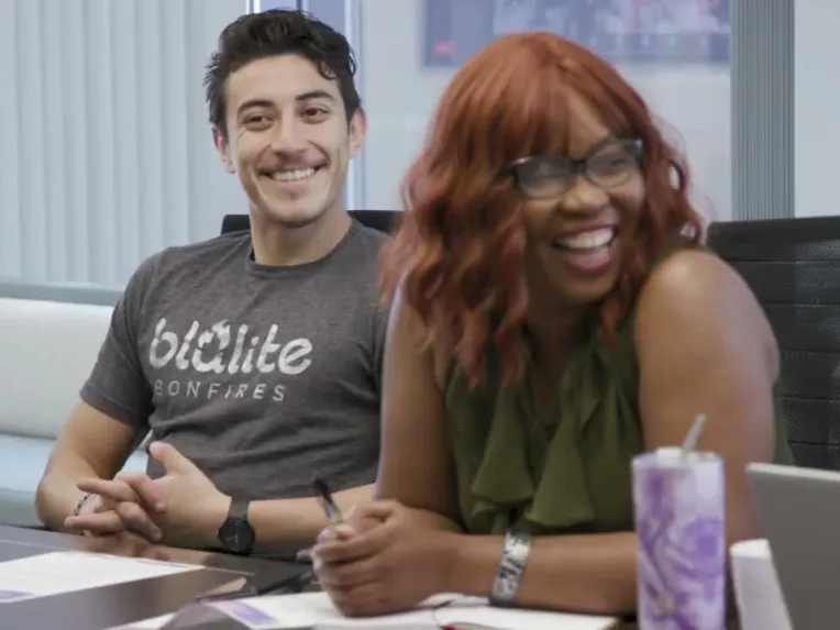 Two people sit at a conference table, smiling and holding pens, with documents, a laptop, and a tumbler in front of them during a meeting.