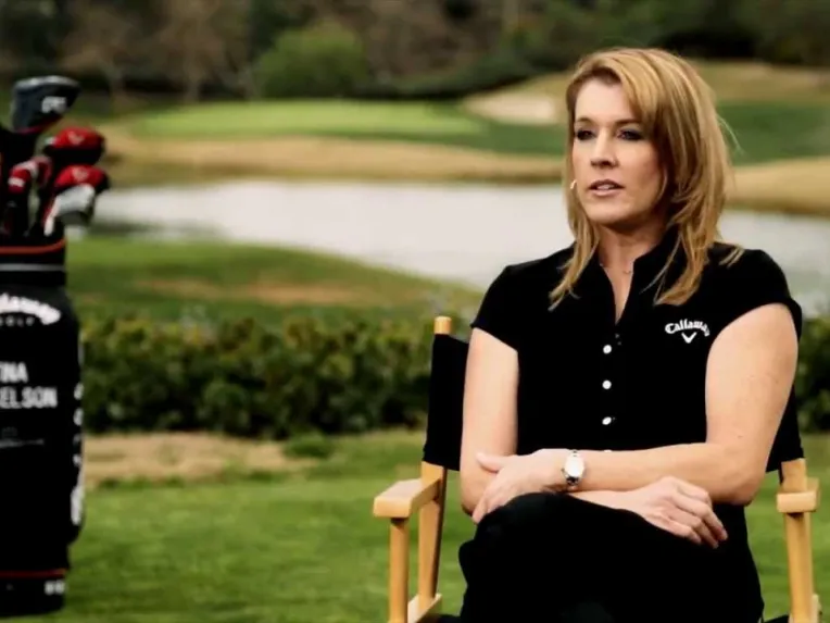 A woman in a black Callaway shirt sits on a chair outdoors by a golf course, with a golf bag labeled "Tina Mickelson" and clubs in the background.