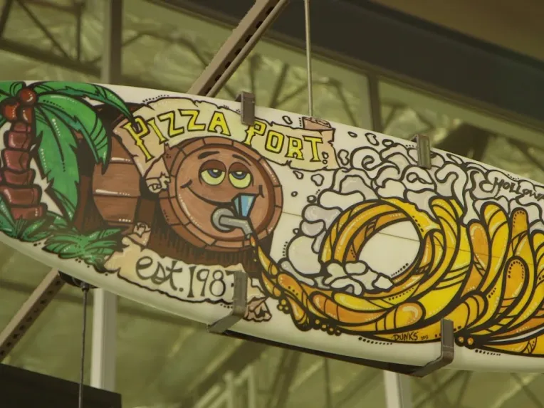 A surfboard decorated with a cartoon design, including a palm tree, a smiling barrel figure, and the text "PIZZA PORT" and "est. 1987," hangs indoors.