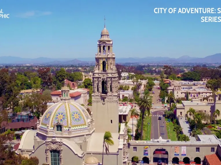 Aerial view of Balboa Park in San Diego featuring historic Spanish-style buildings, lush greenery, and distant mountains under a clear blue sky.