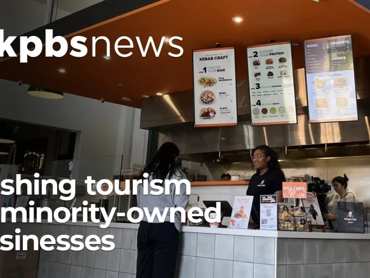 Two women stand at the counter of a fast-casual restaurant as a third person works in the background; kpbs news headline promotes tourism to minority-owned businesses.