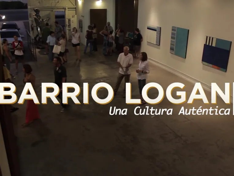 People gather inside an art gallery with modern paintings on the walls. Large text reads “Barrio Logan: Una Cultura Auténtica.”.