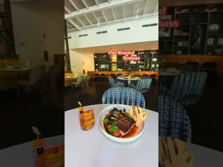A table in a restaurant with a bowl of food, bread slices, and a drink; “Pan Roasted Seabass” sign is visible in the background.