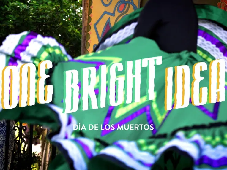 A person in a green folkloric dress dances outdoors; text over the image reads "ONE BRIGHT IDEA" and "Día de los Muertos.