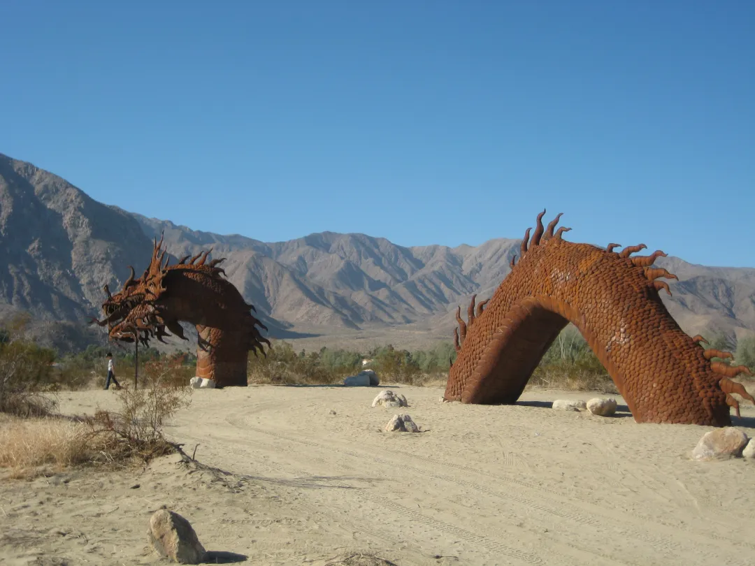 Rusty dragon sculpture arches against a blue sky and mountainous backdrop.
