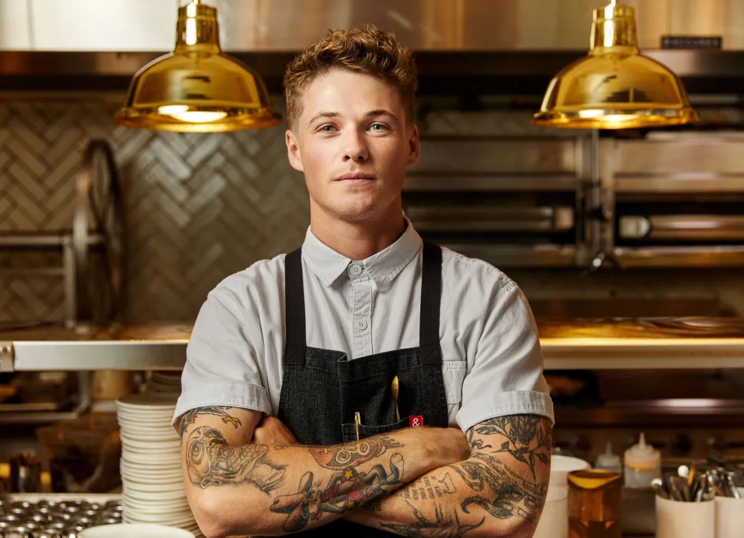 A chef with tattoos crosses arms, standing in a well-lit kitchen with gold pendant lights.