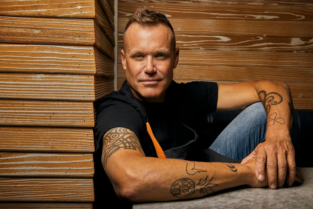 A person with tattoos sitting casually beside wooden panels.