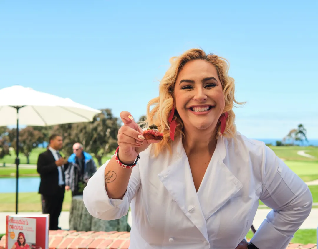 Chef holding a dish outdoors, with a scenic view in the background and people mingling.