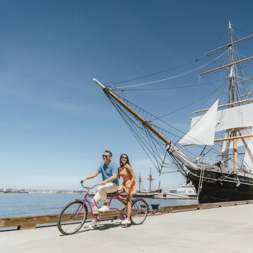 A couple riding a bike on San Diego's Embarcadero with the Star of India at the Maritime Museum in the background