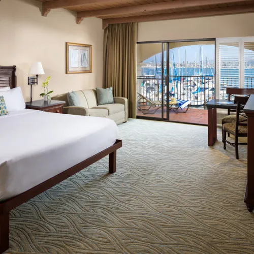 Hotel room with a king bed, sofa, desk, and chairs; sliding glass doors open to a balcony with a view of a marina and boats.