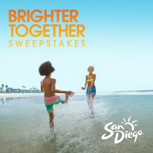 Two children play in the surf at a beach, with palm trees and a clear blue sky in the background.