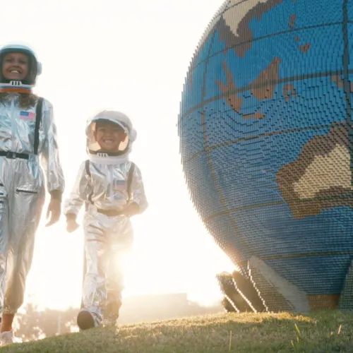 Two child astronauts walk beside a large globe sculpture at sunset.