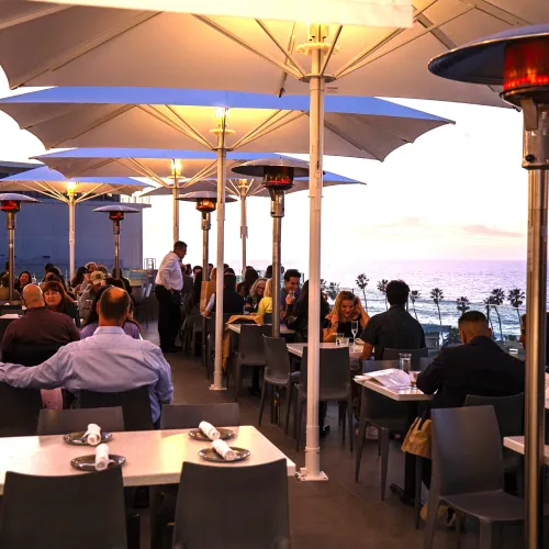The outdoor terrace at George's at the Cove restaurant in the seaside La Jolla neighborhood of San Diego, with diners under large umbrellas, overlooking the ocean at sunset.
