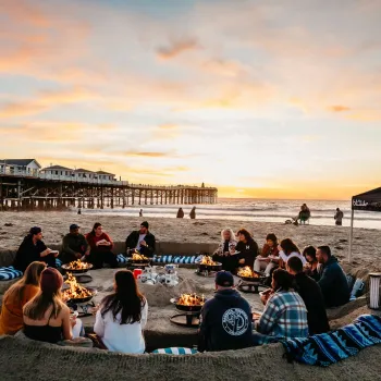 A group of people sit around fire pits in a sand pit on the beach at sunset, with a pier and ocean in the background.