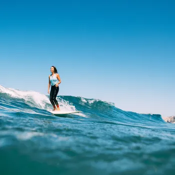A person in a blue top and black wetsuit is surfing on a small wave in the ocean under a clear blue sky in San Diego.