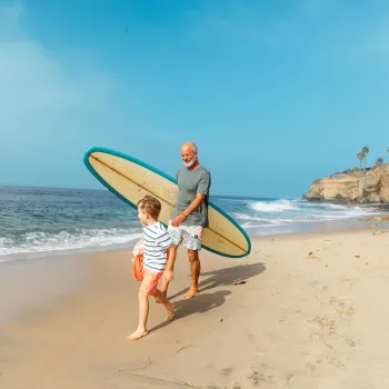 An adult and a child walk barefoot on a sandy beach; the adult carries a surfboard while waves approach the shore in the background.