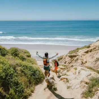 Two people with backpacks walk down a sandy trail in Torrey Pines toward a Blacks Beach, surrounded by greenery and cliffs, with the ocean visible in the background.