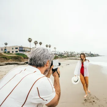 A man takes a photo of a woman posing on a sandy beach near the ocean. The woman holds a hat and smiles, and palm trees and houses are visible in the background.