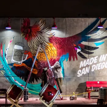 A large, colorful mural of a rooster holding a beer mug with the words "MADE IN SAN DIEGO" painted on a wall, illuminated by overhead lights, with a red bench and drums in front in Convoy, San Diego's Pan Asian Cultural District.