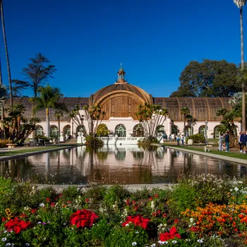 Balboa Park Botanical building with arched windows behind a reflective pond, surrounded by colorful flower beds, palm trees, and visitors on a clear day.