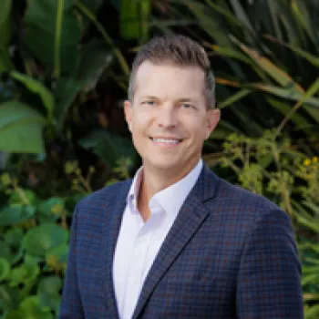 Brian Hilemon smiles in a checked blazer and white shirt while standing outdoors in front of green plants.