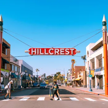 Pedestrians stroll under the Hillcrest sign in Uptown San Diego under a blue sky.