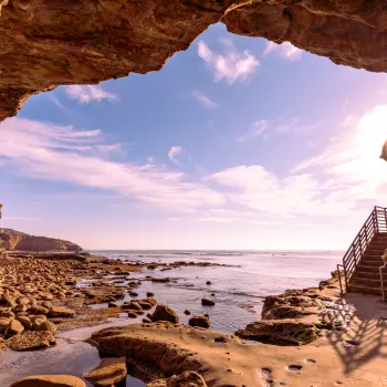 View from inside a rocky seaside cave in Point Loma looking out to the ocean, with a staircase on the right and sunlight streaming through the entrance.