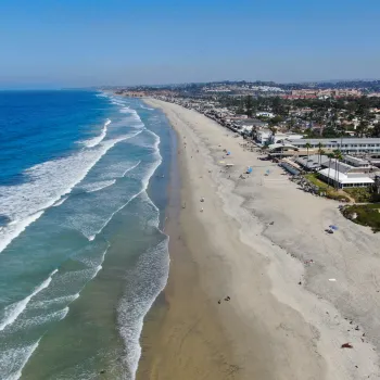 Aerial view of Del Mar's wide sandy beach with waves along the shoreline, people scattered on the sand, and buildings lining the coast under a clear blue sky.