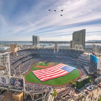 A large American flag is displayed on petco park, with four jets flying in formation overhead and a crowd filling the stands.
