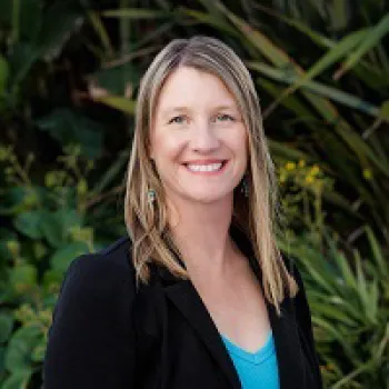 Fiona Jahn with straight blonde hair, wearing a black blazer and blue top, stands smiling outdoors in front of green foliage.