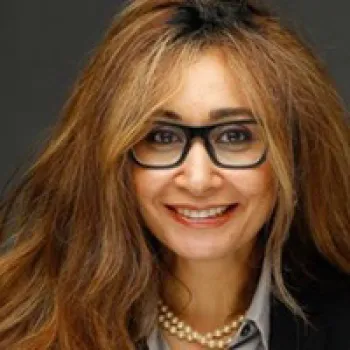 Hanaa Zahran with long wavy brown hair, wearing black glasses. a gray blouse, black blazer, and a pearl necklace, smiling at the camera against a dark background.