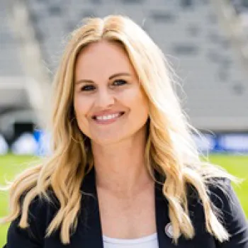 Jennifer Bower with long blonde hair smiles while standing outdoors in a stadium. She is wearing a dark blazer over a white top.
