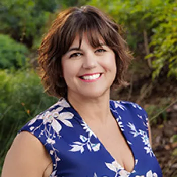 Jen Malan with short brown hair wearing a blue floral dress is smiling outdoors with greenery in the background.