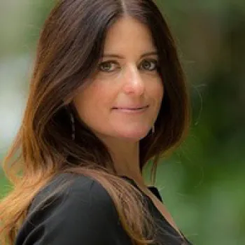 Michelle Metter with long brown hair and a black top poses outdoors, looking at the camera with a slight smile; green blurred background.