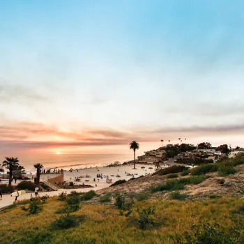 People walk along a path overlooking a Moonlight beach in Encinitas at sunset, with palm trees, parked cars, and houses visible in the background under a partly cloudy sky.