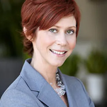 Nancy Bjork with short red hair wearing a gray blazer and necklace, smiling at the camera with a blurred indoor background.