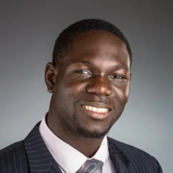 Kazeem Omidiji in a dark pinstripe suit, light pink shirt, and tie smiles at the camera against a plain gray background.