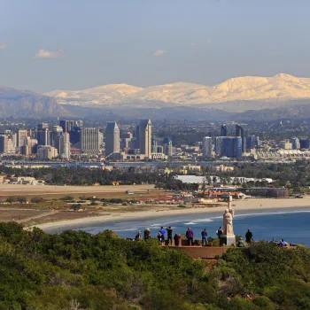 A group of people stands on a hill overlooking the San Diego skyline, with a beach, bay, and distant snow-capped mountains in the background.
