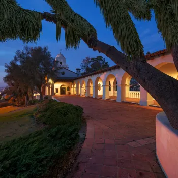 A large tree frames a Spanish-stylePresidio Museum with arches and a dome, illuminated at dusk, overlooking a cityscape with visible lights in the distance.