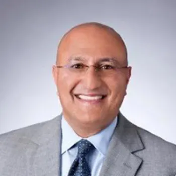Sam Attisha wearing glasses, a light gray suit, blue shirt, and patterned tie poses in front of a plain gray background.